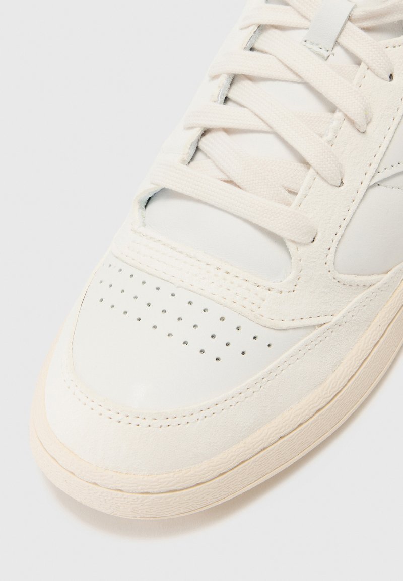 Zapatilla blanca de cuero con acentos de ante texturizado, caja de dedos perforada y cordones planos. Presenta una suela de goma. Diseño limpio y minimalista.