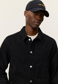 Μαύρο τζιν μπουφάν με υφή λουλουδιών, ασημένια κουμπιά με σούστα και πλαϊνές τσέπες. Φορεμένο με μπλε σκουφάκι με κίτρινο κείμενο.