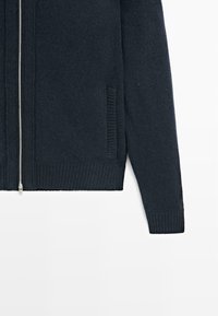 Cardigan zip-up di un blu navy scuro con texture a coste, dettagli a righe verticali sui lati e chiusura con zip in metallo.