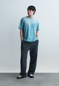 Νέος με μαύρα μαλλιά, που φοράει ανοιχτό γαλάζιο φαρδύ T-shirt, μαύρο φαρδύ παντελόνι, μαύρα και λευκά παπούτσια, στέκεται με τα χέρια στις τσέπες.