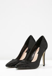 Ett par svarta högklackade kvinnors skor med spetsiga tår, täckta i glänsande paljetter, som står på en vit yta.