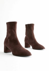 Bottines en daim marron avec un haut tige, une pointe carrée, un talon épais et un détail de fermeture éclair sur le côté. Texture lisse et design minimaliste.