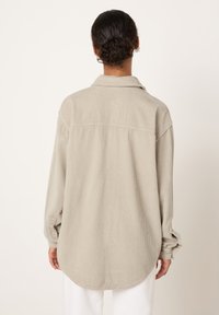 Osoba s tmavými kudrnatými vlasy svázanými do drdolu má na sobě volnou béžovou manšestrovou košili a bílé kalhoty, viděna zezadu na neutrálním pozadí.