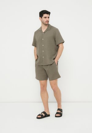 Homme portant une chemise à manches courtes boutonnée vert olive et un short assorti, debout les mains dans les poches, avec des sandales noires à double bride.