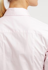 Camicia button-up rosa chiaro con colletto, texture liscia e dettaglio della cucitura a yoke sul retro. Il tessuto appare morbido e leggermente aderente.