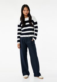 Suéter de punto a rayas en azul marino y blanco con textura acanalada y dobladillo deshilachado, combinado con pantalones navy de corte relajado y zapatos blancos.