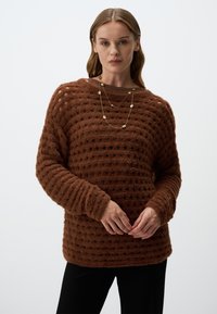 Bruine textuurtrui met een losse pasvorm, voorzien van een stippenpatroon, geribbelde halslijn en lange mouwen. Gecombineerd met een gelaagde gouden ketting.