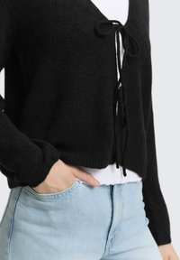 Persoon draagt een zwarte cardigan met knoop aan de voorkant over een wit shirt en lichtblauwe jeans met hoge taille, hand in de zak gestoken.