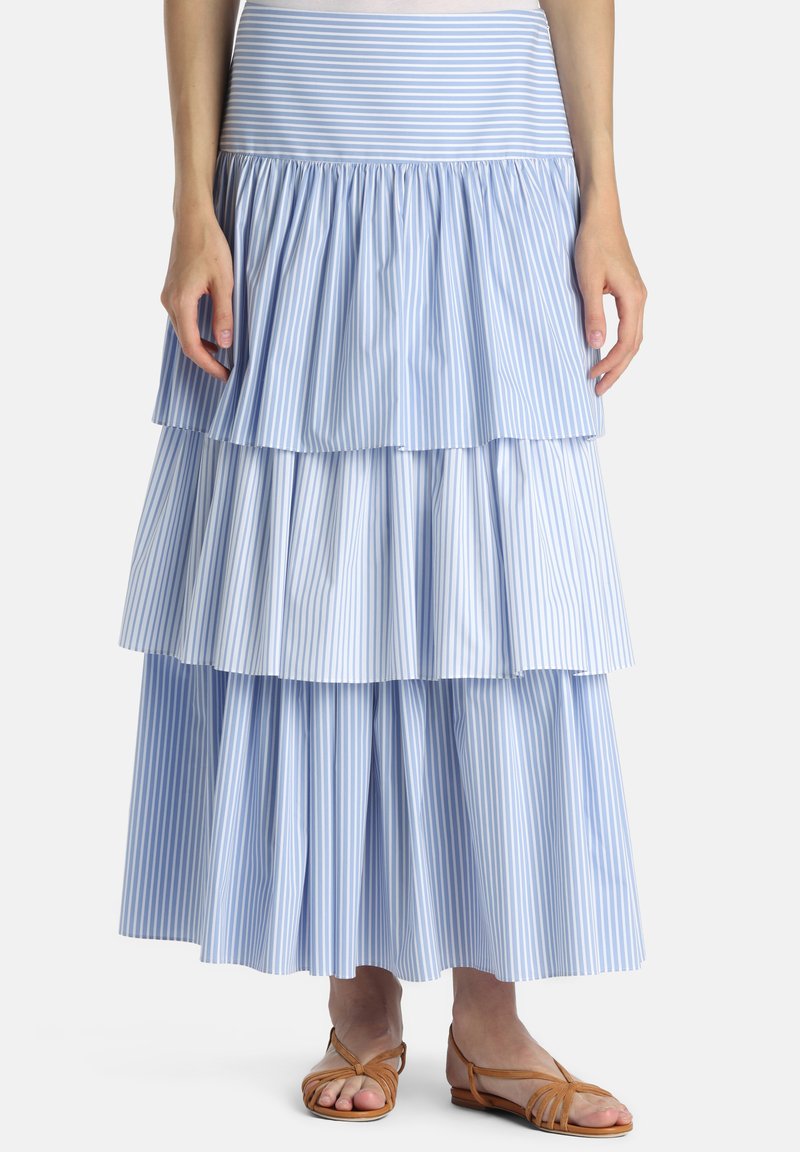 Kvinde iført en blå og hvid, vertikalt striped, lagdelt maxi nederdel med brune flade sandaler på en hvid baggrund.