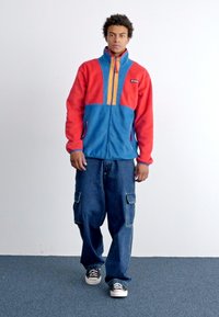 Giacca in pile zip-up rossa e blu con dettagli arancioni, abbinata a jeans blu larghi e scarpe in tela nere. Design semplice e informale.