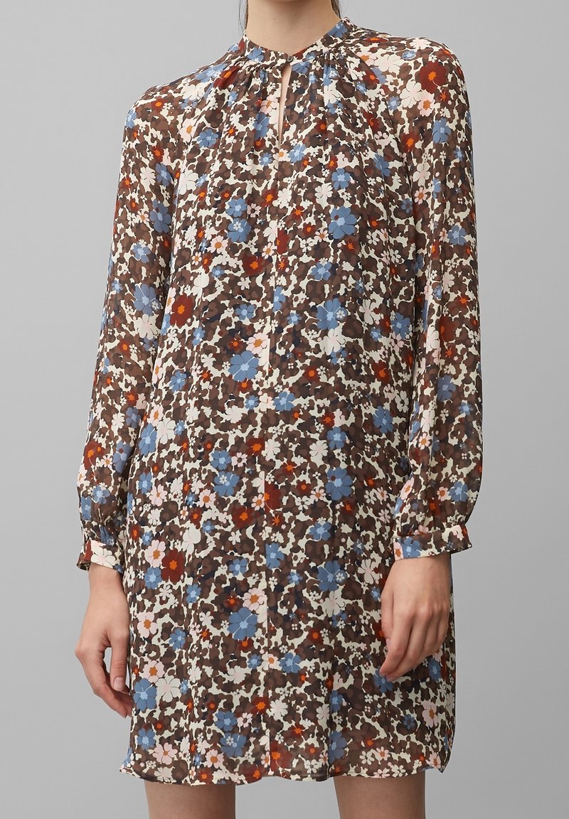 Femme portant une robe à manches longues, longueur genoux, avec un motif floral marron, bleu, blanc et rouge et une petite encolure en forme de trou de serrure.