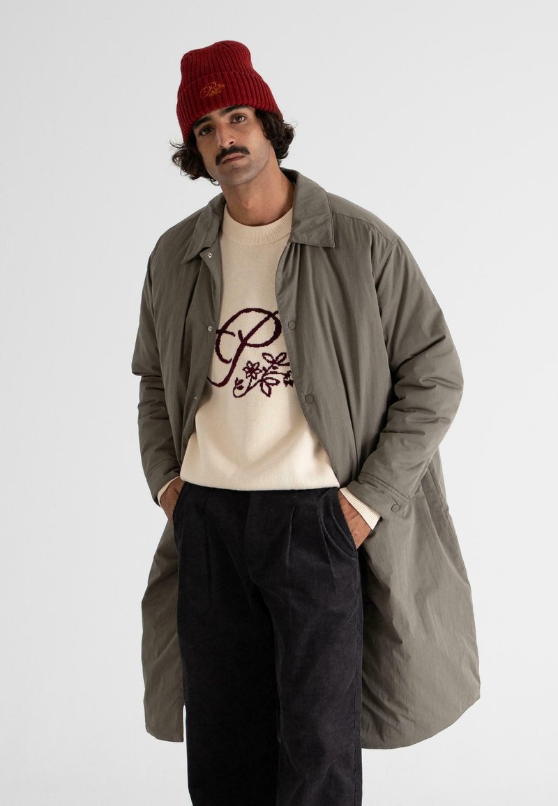 Olivenfarvet frakke med knapper over en creme sweater med burgundy broderi og sorte kordfløjl bukser; rød strikket hue tilføjer kontrast.