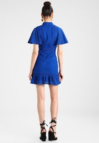 Vestido azul de encaje con mangas cortas y ligeras, dobladillo con volantes y un intrincado diseño recortado. Combinado con sandalias negras de tiras.