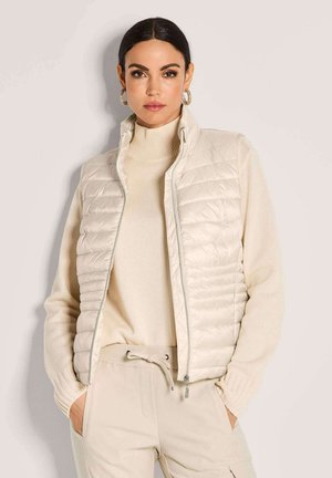 Frau mit dunklen Haaren, die goldene Creolen trägt, eine beige gesteppte Weste über einem cremefarbenen Rollkragenpullover und beige Hosen mit Kordelzug-Taschen.