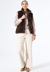 Braune Kunstfellweste mit hohem Kragen und Knopfverschluss, getragen über einem gestreiften Hemd. Kombiniert mit helleren Hosen und braunen Schuhen.