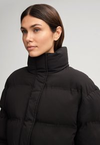 Giacca a vento nera con colletto alto, caratterizzata da un design trapuntato e branding sul tessuto. La texture appare liscia e isolata.