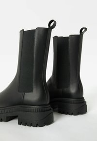 Botas Chelsea de cuero negro con paneles elásticos, suelas de goma gruesa y tiradores en la parte posterior. Textura suave y diseño elegante.