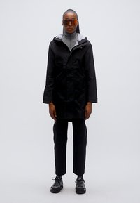 Μαύρο αδιάβροχο μπουφάν με γκρι κουκούλα, φορεμένο πάνω από γκρι πουλόβερ ζιβάγκο, συνδυασμένο με μαύρο παντελόνι και μαύρα αθλητικά παπούτσια. Γυαλιά με πορτοκαλί φακούς.