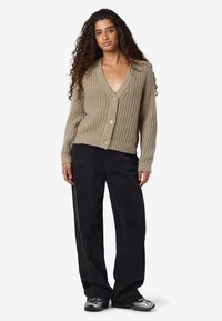 Mulher com cabelo louro encaracolado longo, a usar um cardigan oversized em malha bege, calças pretas largas e sapatilhas prateadas, de pé contra um fundo simples.