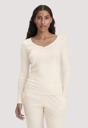 Vrouw met lang donker krullend haar draagt een crèmekleurig langhouwen henley-shirt en bijpassende crèmekleurige broek tegen een effen lichte achtergrond.