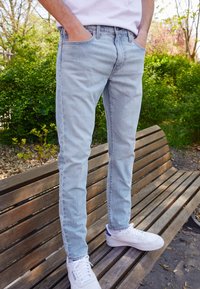 Light blue denim jeans with a slim fit, featuring classic five-pocket styling. Worn with white sneakers, seated on a wooden bench.