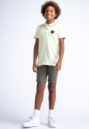 Lichtgele polo met zwarte accenten, voorzien van een logo patch. Gecombineerd met olijfgroene opgerolde shorts en witte sneakers met grijze veters.