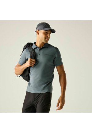 Man wearing a blue polo shirt and cap, carrying a black backpack over one shoulder, looking sideways against a plain background.