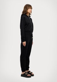 Schwarze Zip-up-Hoodie mit tailliertem Design, kombiniert mit schwarzen Jogginghosen mit Kordelzug. Getragen mit strukturierten schwarzen Sandalen mit Schnallen.