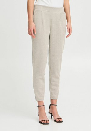 Femme portant un pantalon beige texturé et fuselé ainsi que des sandales à talons noires ouvertes à l'avant avec une bride à la cheville, debout sur fond blanc.