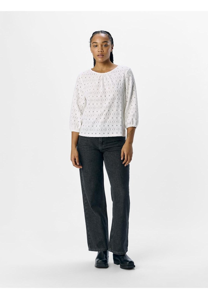 Blusa bianca in pizzo con maniche a tre quarti, scollo rotondo e texture sottile. Abbinata a jeans scuri a vita alta e stivali neri.