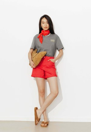 Femme portant un t-shirt gris, un short rouge, un foulard rouge autour du cou, tenant un sac beige et portant des sandales beiges, debout contre un mur blanc.