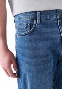 Vaqueros azul de denim con un corte clásico, bolsillos frontales, cierre de botón metálico y detalle de costura en contraste. Textura suave y acabado ligeramente desgastado.