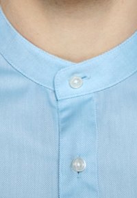 Camicia azzurra chiaro con tessuto testurizzato, caratterizzata da un colletto rotondo e due bottoni bianchi rotondi visibili alla scollatura.