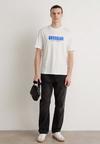 Camiseta blanca con estampado azul "HUGO CREATIVE DEPT", combinada con pantalones de mezclilla negros y zapatillas blancas con suela beige. Bolso negro en la mano.