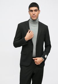 Μαύρο σακάκι με δύο κουμπιά, συνδυασμένο με γκρι τζάκετ με ζιβάγκο. Διαθέτει λοξούς γιακάδες και μια μόνο τσέπη στο στήθος. Λείο υφασμάτινο σχέδιο.