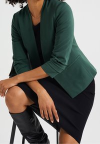 Vihreä bleiseri, joka on valmistettu sileästä kankaasta, ja jossa on huppukaulus sekä kierretyt hihat. Yhdistettynä mustaan ribbimekkoon ja mustiin polvipitkiin saappaisiin.