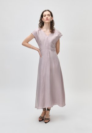 Kobieta z kręconymi włosami, ubrana w jasnofioletową sukienkę bez rękawów z dekoltem w szpic i czarne sandały na paski, stojąca z jedną ręką na biodrze.