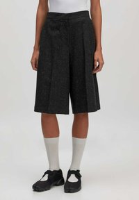 Schwarze, knielange Shorts aus strukturiertem Stoff mit einem gepunkteten Muster, mit gefaltetem Vorderteil und seitlichen Taschen. Getragen mit weißen Socken.