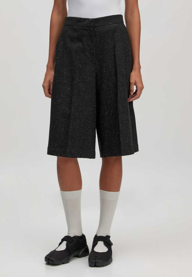 Schwarze, knielange Shorts aus strukturiertem Stoff mit einem gepunkteten Muster, mit gefaltetem Vorderteil und seitlichen Taschen. Getragen mit weißen Socken.