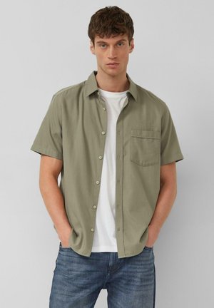 Jeune homme aux cheveux bruns courts portant une chemise olive à manches courtes ouverte sur un T-shirt blanc et un jean bleu, les mains dans les poches.