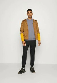 Άνδρας που στέκεται με καφέ σακάκι με κίτρινες μανσέτες, γκρι πουκάμισο, μαύρα παντελόνια και μαύρα αθλητικά παπούτσια, μπροστά σε απλό φωτεινό φόντο.