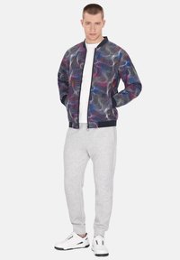 Kleurvolle bomberjack met een draaiend abstract patroon in tinten roze, paars en blauw, gecombineerd met grijze joggingpakken en witte sportieve sneakers.