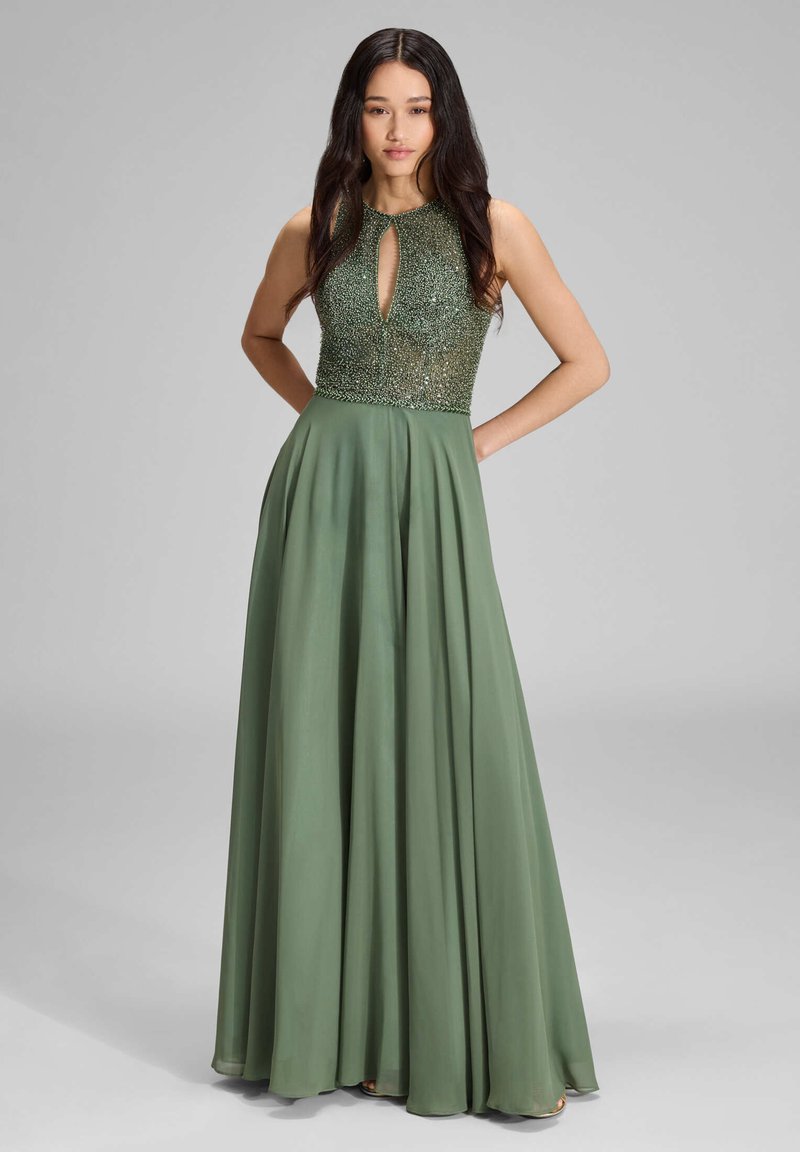 Femme aux longs cheveux noirs portant une robe sans manches vert olive avec un corsage orné de perles et un décolleté en trou de serrure, debout les mains derrière le dos.