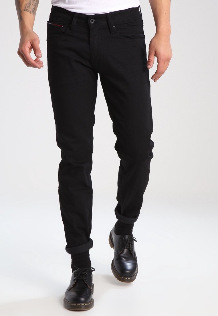 Vaqueros de denim negro con un corte ajustado, diseño de cinco bolsillos y puños enrollados. La tela tiene una textura suave y un minimalismo en los detalles. Llevados con zapatos negros.
