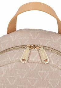 Mochila de cuero rosa claro con superficie texturizada, con herrajes dorados, cierre de doble cremallera y un patrón de logotipos en relieve.