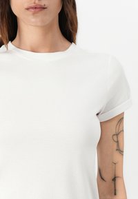 White short-sleeve t-shirt with a rounded neckline, featuring a soft, smooth texture and rolled sleeves. Tattoo visible on the left arm.