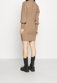 Stickad, figursydd beige klänning med ribbad textur, hög halsringning och puffärmar; kombinerad med svarta ankelstövlar mot en enkel bakgrund.