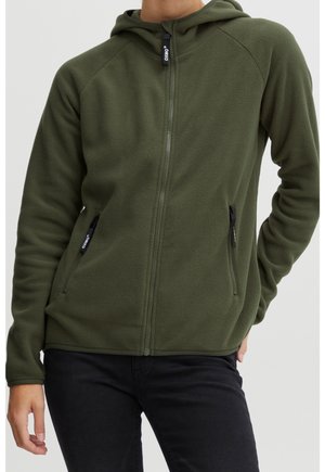 Personne portant un sweat à capuche polaire vert olive zippé avec des poches latérales zippées et un pantalon noir, debout avec une main dans la poche.