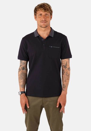 T-shirt polo noir avec une patte de boutonnage à deux boutons, col gris contrastant et détail de poche. Le modèle porte un pantalon vert olive ; des tatouages visibles sur les bras.