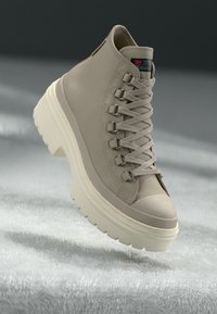 Beige Mid-Cut Sneaker mit Obermaterial aus glattem Leder, strukturiertem Gummisohle, hervorstehenden Metallösen und flachen Schnürsenkeln, mit leicht erhöhtem Absatz.